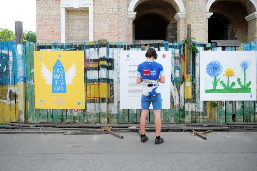 На заборе Гостиного Двора устроили выставку репродукций детских рисунков «Сидимолітаємо»